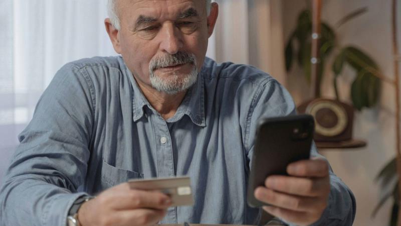 An older man using a smartphone while looking at a bank card.